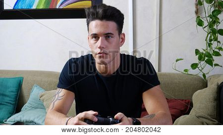 Attractive young man using joystick or joypad for videogames, sitting on couch at home in his living room, staring at TV screen