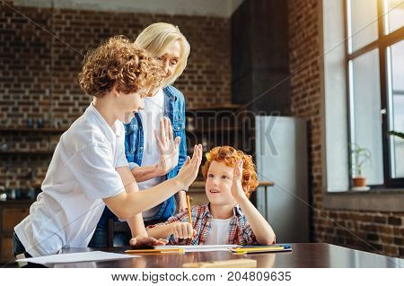 One for all and all for one. Adorable ginger haired boy giving a high fie to his granny and older brother while sitting at a table and drawing with colorful pencils.