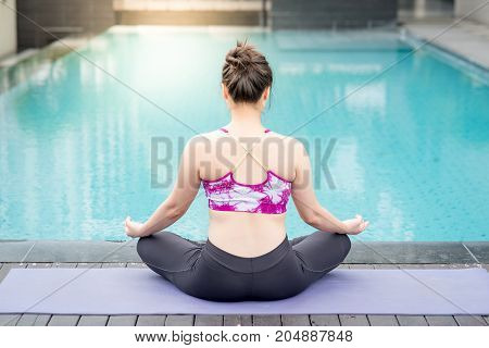 Beautiful young Asian woman doing yoga exercise with lotus position posing near swimming pool. Healthy lifestyle and good wellness concepts