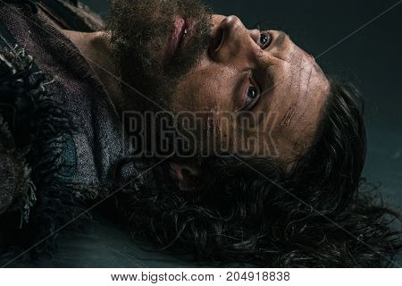 Halloween zombie lying on paving asphalt. Vampire hipster with red blood outdoors. Holiday celebration concept. Man with bloody beard and hair. War soldier with wounds on face.