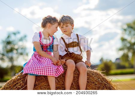 Two kids in traditional Bavarian costumes in wheat field. German children sitting on hay bale during Oktoberfest. Boy and girl play at hay bales during summer harvest time in Germany. Best friends
