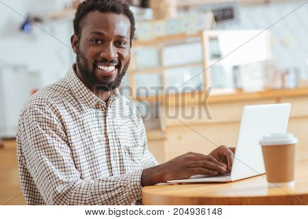 Business correspondence. Charming smiling man sitting at the table in the cafe and typing an e-mail while smiling at the camera