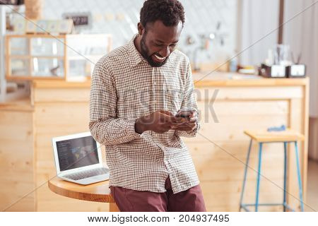 Mood-boosting chats. Cheerful pleasant young man leaning on the table in the cafe and sending text messages to his friends via his phone while smiling happily