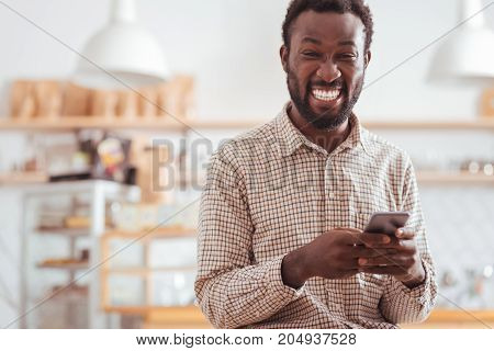 Hilarious messages. Overjoyed upbeat young man standing in the coffee house and laughing hard while reading funny text messages from his friends on his phone