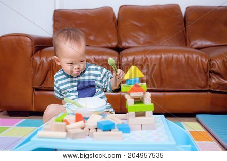 Cute Asian 20 months / 1 year old toddler baby boy child play with colorful wooden blocks. Kid playing with educational toys while eating snack in living room at home photo in real life interior