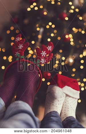 Detail of male and female feet wearing warm winter socks with small Santa's hat and antlers placed on the table with Christmas tree and Christmas lights in background. Selective focus on the antlers
