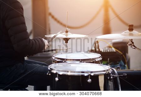 A Street Musician, Wearing A Jacket And Jeans, Plays With Battered Drumsticks On A Drum Set.