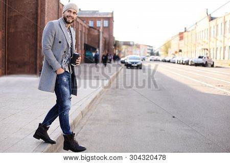 Handsome Young Man In Grey Coat And Hat Crossing The Street With A Cup Of Coffee.