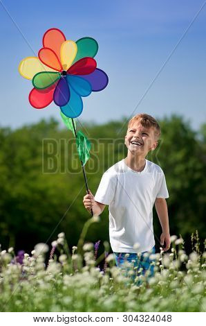 Happy Little Boy With Colorful Weathercock Walking On A Meadow In Sunny Day. Child Playing Windmill 