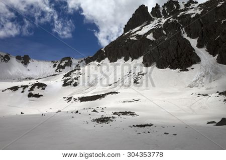 High Sunlit Mountains With Snow Cornice And Avalanche Track, Snowy Plateau And Two Small Silhouettes