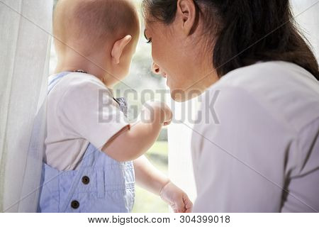 Young Woman And Her Little Son Looking At The Street Throgh The Window At Home