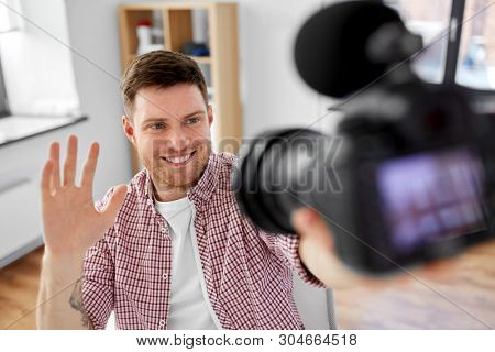 blogging, videoblog and people concept - smiling male video blogger with camera videoblogging and waving hand at home office