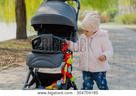 A Little Girl Is Rolling Baby Carriage In The Park. Child In The Park Playing With Pram.