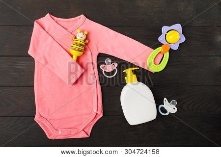 Pink Bodusuit Holding Two Rattles And Two Sylicone Pacifiers With Baby Soap On Brown Wooden Backgrou