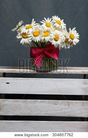 A Bouquet Of Field Daisies In A Glass Vase. On The  Wooden Box. Genus Of Perennial Flowering Plants