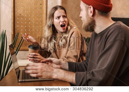 Image of a shocked quarrel young loving couple in cafe using laptop computer drinking coffee.