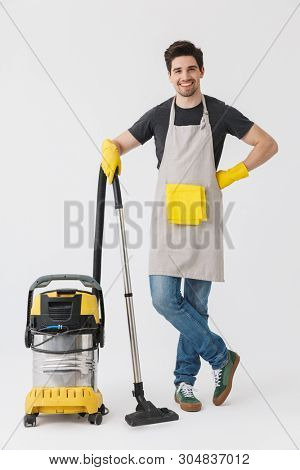 Full length of a handsome brunette houseman wearing apron standing isolated over white background, cleaning with vacuum cleaner