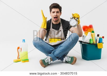 Handsome brunette houseman wearing apron sitting with bucket full of detergents isolated over white background, holding alarm clock