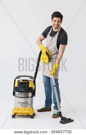 Full length of a handsome brunette houseman wearing apron standing isolated over white background, cleaning with vacuum cleaner