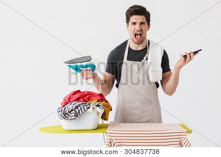 Handsome angry brunette houseman wearing apron standing isolated over white background, ironing clothes on a board, talking on mobile phone