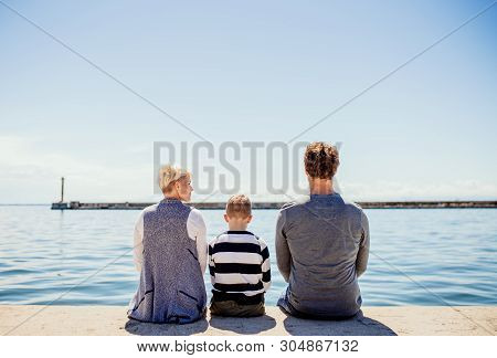 Rear View Of Young Family With Son Sitting On Conrete Pier Outdoors By The Sea.