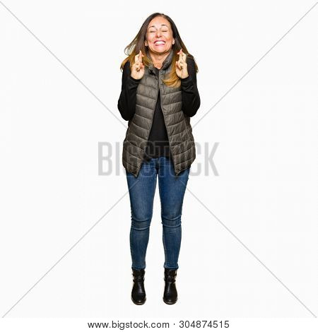 Beautiful middle age woman wearing winter vest smiling crossing fingers with hope and eyes closed. Luck and superstitious concept.