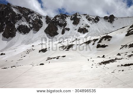 High Mountains With Snow Cornice And Avalanche Trail, Snowy Plateau And Two Small Silhouette Of Hike