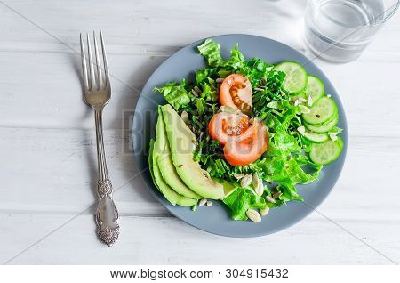 Fresh  Green Salad With Cucumber, Lettuce, Avocado And Tomatoes And Glass Water On A White Wooden Ta