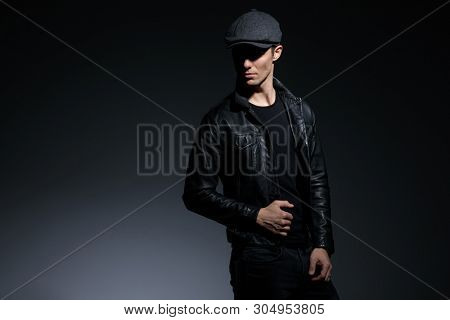 Dramatic looking guy pulling his black leather jacket and confidently looking forward while wearing a hat and jeans, standing on gray studio background