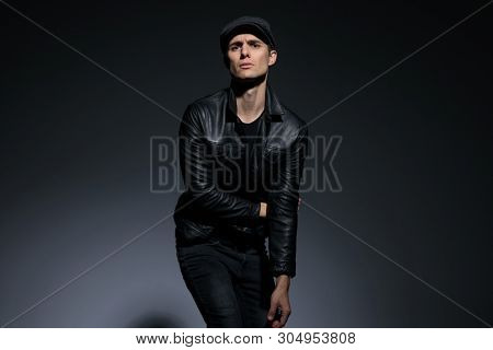 Artistic man posing as powerless and looking to the camera while wearing a cap and black leather jacket, standing on gray studio background