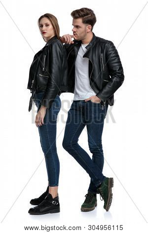 Curious boyfriend holding his hand on his chin, looking to the side and leaning on his girlfriend, both wearing black leather jackets and jeans, standing on white studio background
