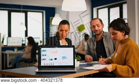 Team Of Colleagues Employees Cooperating Sitting At Desk In Startup Company Office Analysing Graphs,