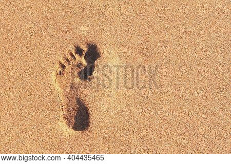 One Footprint Of A Human Barefoot Left Foot, Imprinted In Close-up On Smooth Brown Sand Bathed In Su