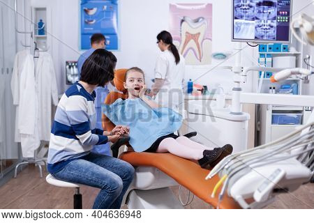Little Girl In Dentist Office Touching Mouth With Hand And Painfull Expression. Child With Her Mothe
