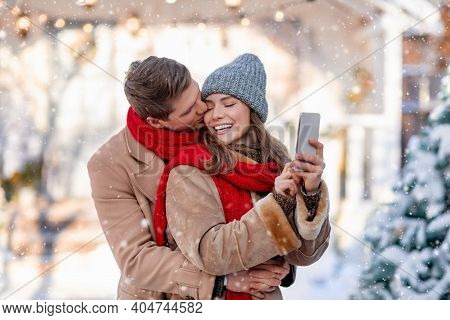 Young Man And Woman Hugging And Taking Selfie On Mobile Phone On Sowy And Sunny Winter Day, Sharing 