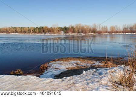 Section Of Wide Plain River Partially Covered With Thin Ice Against The Clear Sky And Trees On Oppos