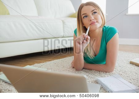 Thoughtful blonde lying on floor doing her assigment using laptop in living room at home