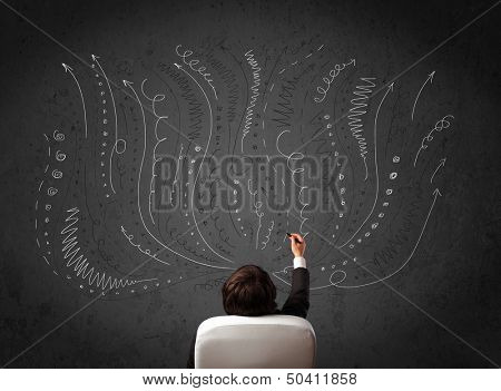Thoughtful young businessman sitting and deciding in front of a chalkboard with sketched arrows and lines in different directions