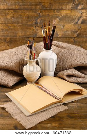 Composition with colorful watercolors, brushes in vase and sketcher on wooden background 