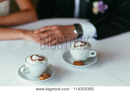 Bride's And Groom's Hands Holding Each Other On A Table With Two Capiccino