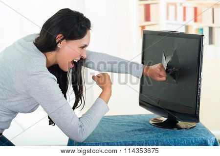 Angry female brunette punching computer screen with large hole on it