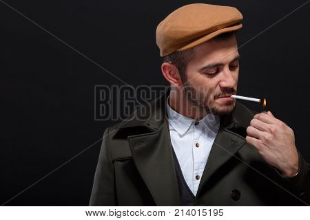 A man on a black background, retro style, in a cap lights a cigarette, screwing up his eyes.