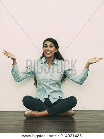 Portrait of smiling at camera middle-aged woman sitting on floor with crossed legs and outstretched hands with white wall in background. Front view.