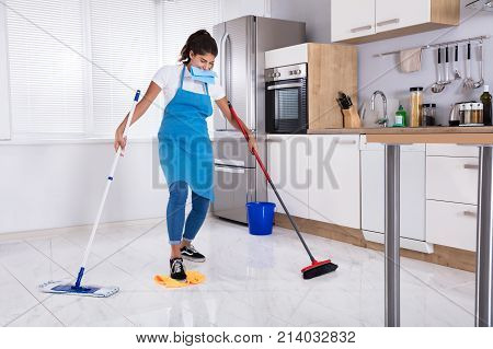 Young Female Janitor Multitasking By Sweeping And Mopping In Kitchen At Home