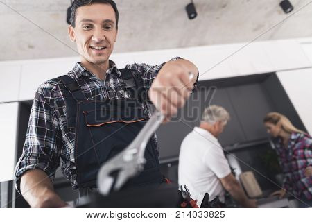The plumber is posing in the kitchen. He holds a wrench in his hands and smiles. Behind him is a client and communicates with the second plumber. A black toolbox is next to it.