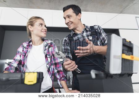 A man and a woman plumber will be cheated in the kitchen. They have just successfully repaired the sink for the customer. Next to them is a black box with a tool.