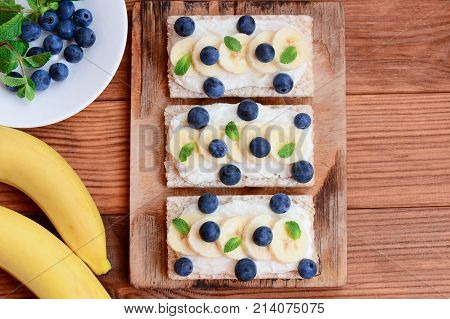 Cottage cheese, fresh bananas and berries sandwiches with crisp bread on wooden board. Vegetarian sandwiches recipe. Top view