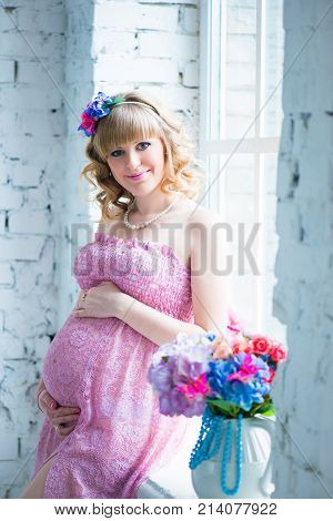 Pregnant woman smiling, looking at belly, holding flowers. Pink dress