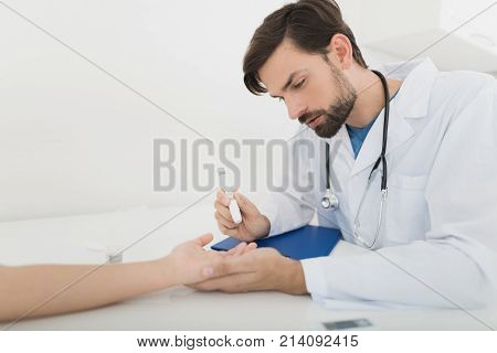 The doctor takes a blood sample from the boy to check it for sugar. The boy patiently endures the procedure. The doctor holds a scarifier in his hands. They are in a modern clinic.