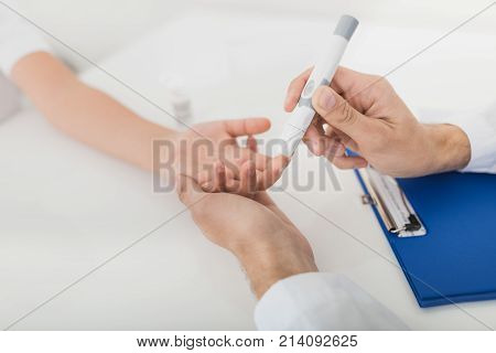 The doctor takes a blood sample from the boy to check it for sugar. The boy patiently endures the procedure. The doctor holds a scarifier in his hands. They are in a modern clinic.